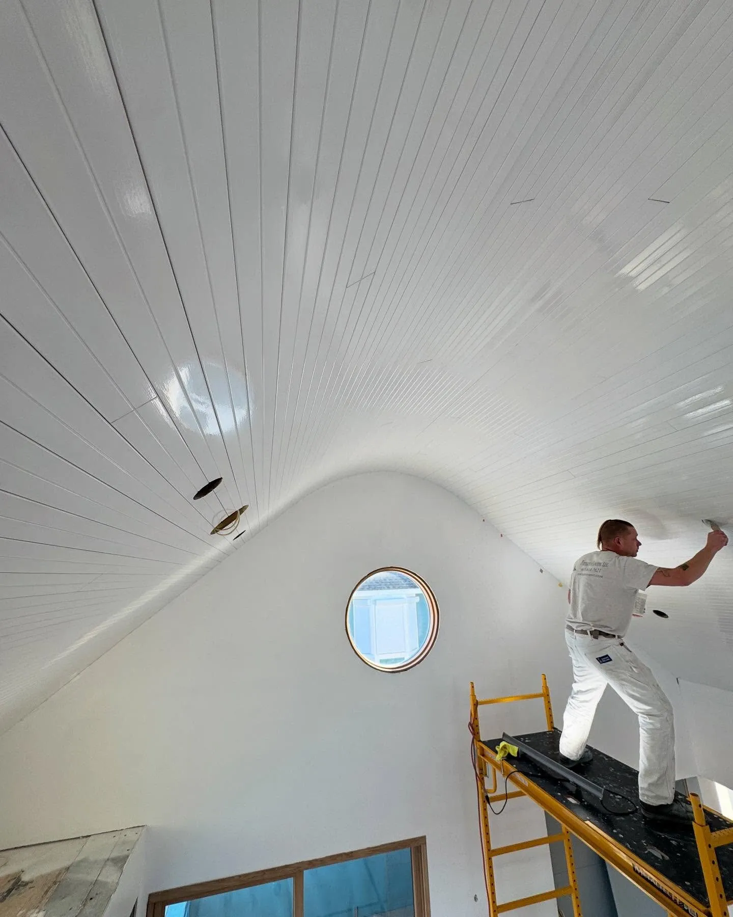 Painter on scaffolding finishing a curved white shiplap ceiling with a porthole window