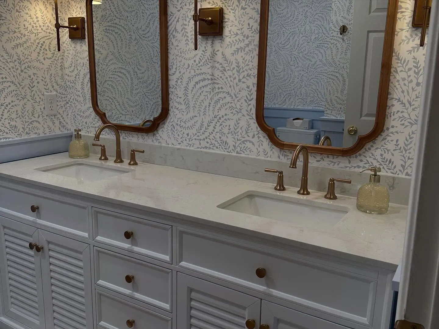 Bathroom with leafy blue-and-white wallpaper above a double vanity and brass mirrors
